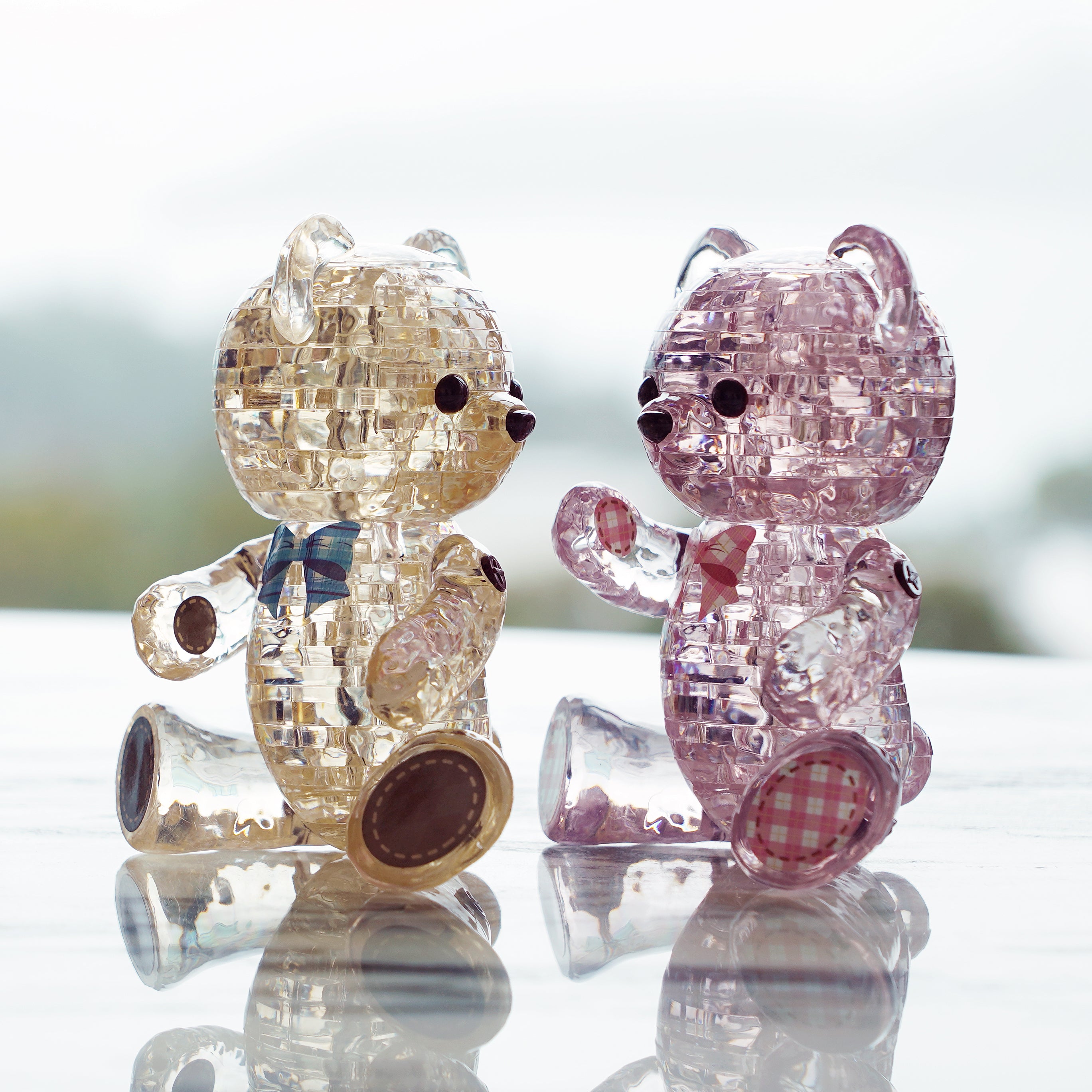 Off-white colored Jewel Bear Henry and pink colored Jewel Bear Lily sitting on a glass table under natural lighting. 